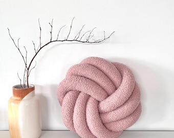 May include: A dusty pink, circular knot pillow made of textured, fuzzy fabric. Next to it stands a ceramic vase with a white and tan glaze, containing a single, thin, dark brown leafless branch with several small, delicate twigs.