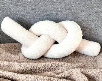 May include: A white fluffy knotted pillow on a brown fuzzy blanket. The pillow is shaped like a knot and is made of a soft, plush material.