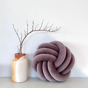May include: A large, round, purple velvet cushion tied in a knot. The cushion is sitting on a white surface next to a vase with a brown and white design.