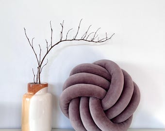 May include: A large, round, purple velvet pillow tied in a knot. The pillow is sitting on a white surface next to a vase with a brown and white design.