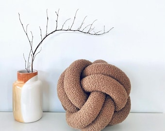 May include: A brown, knotted decorative pillow with a textured surface. A white and orange vase with a branch is to the left of the pillow. The pillow is a unique home decor item.