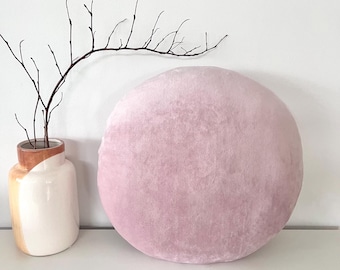 May include: A round, soft-textured pillow in a dusty rose pink color. Beside the pillow sits a ceramic vase with a white and tan color-blocked finish, containing a single thin, dark brown branch with small, delicate twigs extending outward.