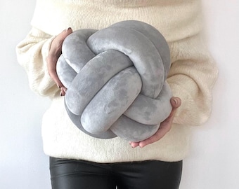 May include: A soft, light gray velvet knot pillow shaped like a sphere with interconnected fabric loops. The surface of the pillow has a smooth, plush texture and a consistent matte finish, creating a rounded, sculptural form that is held in place.