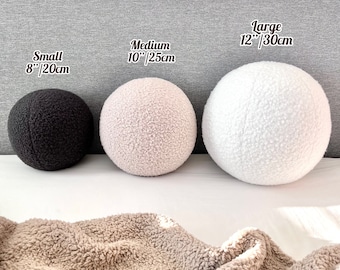 May include: Three round fluffy pillows in different sizes. The smallest pillow is black, the medium pillow is light pink, and the largest pillow is white. The pillows are on a gray textured surface with a tan fuzzy blanket in the foreground.