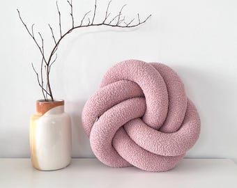 May include: A soft, pink knot-shaped decorative pillow. The pillow is made of a textured fabric and sits on a white surface. A small vase with a branch is to the left of the pillow.
