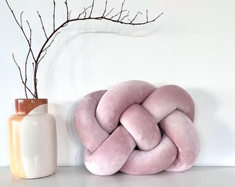 May include: A pink knotted pillow with a soft, velvety texture. The pillow is in the shape of a square with rounded corners and is made of multiple knots.