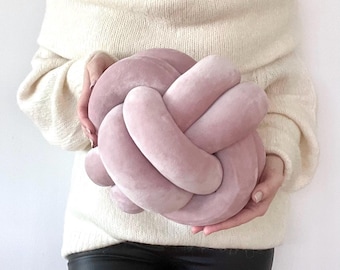 May include: A dusty rose pink knot pillow featuring a continuous, interconnected tubular design. The surface of the pillow has a soft, velvet-like texture, and the overall shape is spherical, formed by multiple loops of fabric woven into a tight knot.