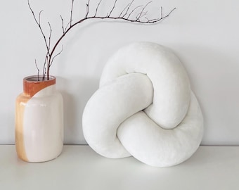 May include: A white knot-shaped pillow with a smooth texture positioned next to a ceramic vase. The vase features a cream-colored base with a tan and orange painted upper section, containing a single thin, dark brown, leafless branch.