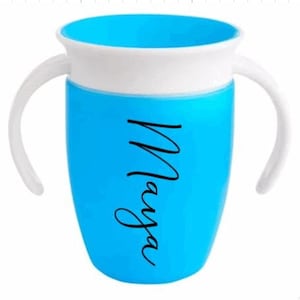 Puede incluir: Un vaso de entrenamiento azul y blanco con dos asas blancas. La taza tiene el nombre "Maya" en escritura negra en el lateral. El interior de la taza es azul.