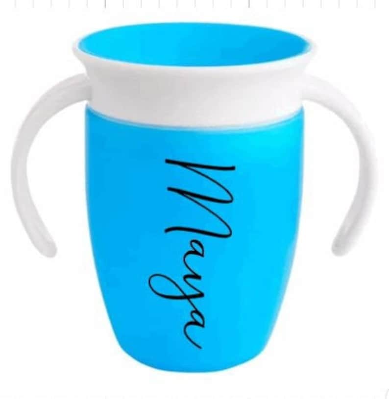 Puede incluir: Un vaso de entrenamiento azul y blanco con dos asas blancas. La taza tiene el nombre "Maya" en escritura negra en el lateral. El interior de la taza es azul.