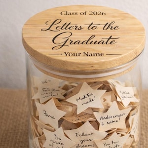 May include: A glass jar with a wooden lid, labeled "Class of 2026 Letters to the Graduate." The jar is filled with star-shaped notes. The words "Graduation Gift" are displayed on the right.