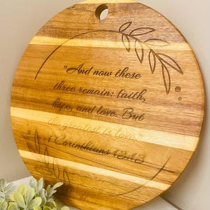 May include: Round wooden cutting board with a light and dark wood grain pattern. The board features engraved text from 1 Corinthians 13:13, along with a decorative leaf design. A hole is drilled at the top for hanging.