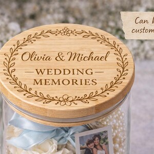 May include: A glass jar with a wooden lid engraved with "Olivia & Michael - WEDDING MEMORIES" and a floral design. The jar contains white flowers, a photo, and a blue ribbon. Text on a tag reads "Can be customized".
