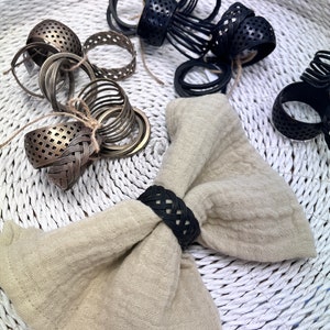 Può includere: Un set di anelli per tovaglioli in metallo nero e oro. Gli anelli sono intrecciati e hanno un design geometrico. Gli anelli sono esposti su una superficie di vimini bianca con un tovagliolo di lino beige.