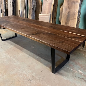 Puede incluir: Una mesa larga de madera marrón oscura con una tapa de borde vivo y patas de metal negro. La mesa está hecha de madera de nogal y tiene un acabado natural.