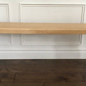 Peut inclure: Un banc en bois naturel avec une finition brun clair et un design simple. Le banc a un dessus rectangulaire et deux ensembles de pieds en bois.