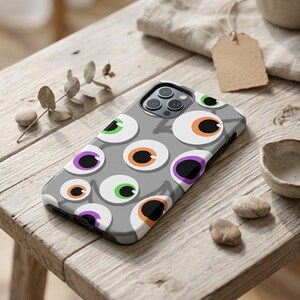 May include: A gray smartphone case featuring a repeating pattern of cartoon eyes with white sclera and irises in shades of orange, green, and purple. The case has a cutout for a triple-lens camera system on the back and covers the device edges.