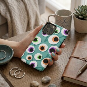 May include: A teal smartphone case featuring a repeating pattern of cartoon eyes with white sclera and irises in shades of green, orange, and purple. The case has a cutout for a triple-lens camera system and is held in a hand against a desk.
