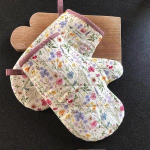 Può includere: Un paio di guanti da forno trapuntati con un motivo floreale di fiori rosa, blu, gialli e viola su sfondo crema. I guanti hanno un bordo malva e un anello per appenderli, appoggiati su un tagliere di legno.