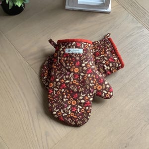 May include: A pair of brown oven mitts with a red trim and a small white tag that says "handmade". The mitts are covered in a pattern of autumn leaves, acorns, and mushrooms.