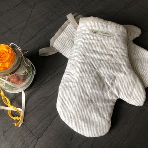 Può includere: Un paio di guanti da forno bianchi con strisce grigie. I guanti sono trapuntati e hanno un anello per appenderli. I guanti hanno un'etichetta che dice "Handmade" e un indirizzo del sito web.