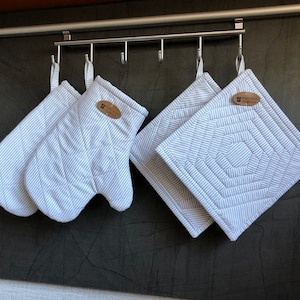 Può includere: Un set di guanti da forno e presine a righe bianche e azzurre appesi a una barra di metallo. I guanti da forno e le presine hanno un design trapuntato e una piccola etichetta marrone.