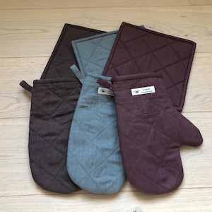 Puede incluir: Conjunto de cuatro agarraderas acolchadas en marrón y azul. Dos agarraderas tienen forma de guantes de horno y dos son cuadradas. Las agarraderas tienen un patrón de diamante y el texto "remarkable" está impreso en una de las agarraderas.
