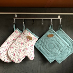 Può includere: Due guanti da forno con stampa floreale e due presine imbottite blu sono appesi a un rack argentato. Le presine hanno un motivo geometrico e un piccolo cartellino marrone con del testo.