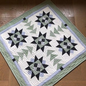 Op de afbeelding: Een handgemaakte quilt met een sterrenpatroon in zwart, wit en tinten groen en blauw. De quilt heeft een witte achtergrond met ster- en driehoekontwerpen. De rand is lichtgroen met een blauwe binnenrand.