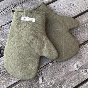 Op de afbeelding: Twee olijfgroene gewatteerde ovenwanten. De wanten zijn gemaakt van een textuurstof en hebben een ruitpatroon. Een klein wit label met tekst is op een van de wanten genaaid. De wanten zijn ontworpen voor gebruik in de keuken.