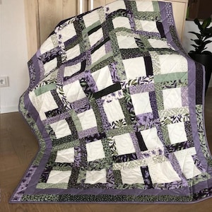 Può includere: Una trapunta fatta a mano con un design patchwork. La trapunta presenta un motivo di quadrati bianchi incorniciati da strisce di tessuto in tonalità di viola, verde e nero, con stampe floreali e di foglie. Il bordo è viola unito.
