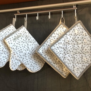 Può includere: Set di quattro presine da cucina bianche e grigie con un motivo maculato. Le presine sono appese a ganci.