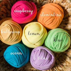 May include: A wicker basket filled with colorful fabric rolls, labeled with names like "raspberry", "clementine", "buttercup", "lemon", "kiwi", "ocean", "lilac".