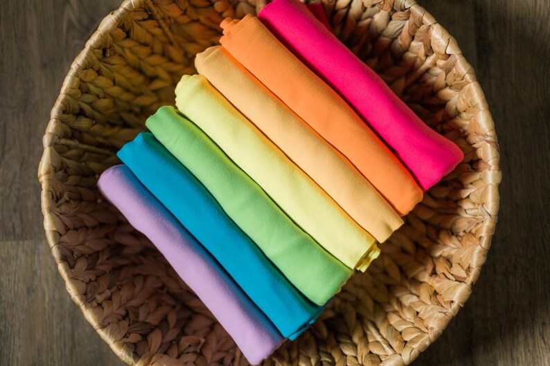 May include: A stack of colorful fabric swatches in a woven basket. The swatches are arranged in a rainbow pattern, starting with purple and ending with red.