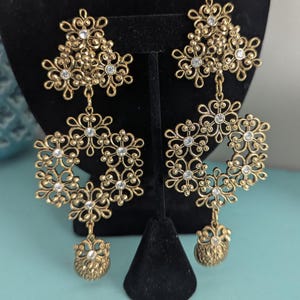 May include: Gold-toned, ornate earrings with a floral design. Each earring features a cluster of small flowers at the top, a circular floral element in the middle, and a small, decorative pendant at the bottom. Clear crystals are set within the floral patterns.