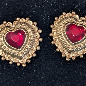 May include: A pair of heart-shaped earrings. Each earring features a gold-colored filigree design surrounding a heart-shaped, deep red gemstone. The earrings are set against a dark background.