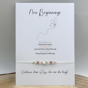 Könnte beinhalten: Eine weiße Präsentationskarte mit dem Text New Beginnings, einem Schmetterling als Strichzeichnung und deutschem Text zu Kristalleigenschaften. Ein zartes weißes Schnurarmband mit klaren, pfirsichfarbenen und weißen Perlen ist mittig befestigt.