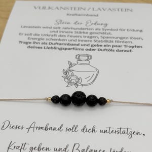 Könnte beinhalten: Ein Armband aus schwarzen Lavasteinen mit goldenen Akzenten. Das Armband ist auf einem neutralfarbenen Kordelband aufgefädelt und wird auf einer Karte mit deutschem Text präsentiert. Die Karte enthält Text über die Eigenschaften des Armbands.