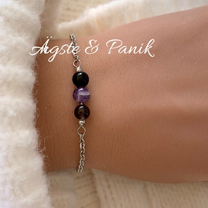 May include: A silver chain bracelet with three beads: a black bead, a purple and white striped bead, and a brown bead. The bracelet is on a person's wrist. The text "Ägste & Panik" is visible in the background.