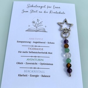 Op de afbeelding: Een zilverkleurige sleutelhanger met een stervormige karabijnhaak en een verticale streng van zes ronde edelsteenkralen in paars, bruin, groen en transparant, bevestigd op een witte kaart met tekst en een lijntekening van een boek en bloemen.