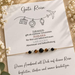 Könnte beinhalten: Ein zartes Armband mit schwarzen, braunen und grauen Perlen an einer dünnen Schnur, präsentiert auf einer weißen Karte mit dem Text 'Gute Reise', Illustrationen von Koffer, Globus, Heißluftballon und Flugzeug sowie Beschreibungen der Steinwirkungen.