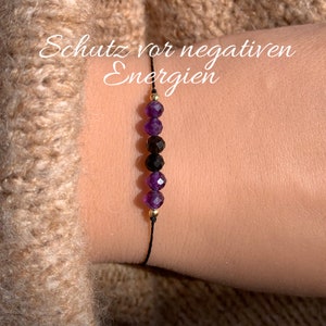 Könnte beinhalten: Ein schwarzes Armband aus Schnur mit Amethyst- und schwarzen Perlen. Das Armband hat goldene Akzente und der Text "Schutz vor negativen Energien" ist im Hintergrund geschrieben.