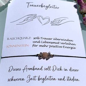 May include: A black string bracelet with a smoky quartz and sunstone charm. The bracelet is accompanied by a card that reads "Trauerbegleiter" and "This bracelet should accompany you and strengthen you in this difficult time."