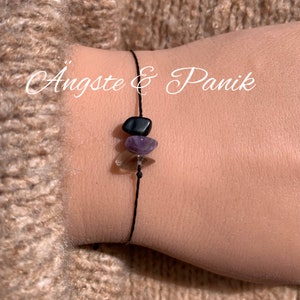 May include: A black string bracelet with three gemstone beads: black, purple, and clear. The bracelet has the text "Ängste & Panik" written on it.