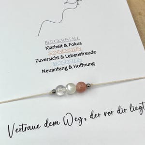 Könnte beinhalten: Ein zartes Armband mit drei kleinen Perlen, einem Bergkristall, einem Mondstein und einem Sonnenstein, auf einem dünnen beigen Band. Das Armband ist auf einer weißen Karte mit deutschem Text und einer Strichzeichnung eines Gesichts befestigt.