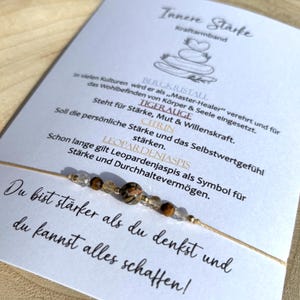 Pode incluir: Uma pulseira de contas com um cordão bege, com contas transparentes, castanhas e estampadas. A pulseira é apresentada num cartão branco com texto alemão sobre força e pedras preciosas. O cartão contém uma citação.