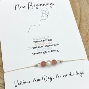 Könnte beinhalten: Ein zartes Armband mit fünf kleinen Perlen in transparenten, pfirsichfarbenen und weißen Tönen an einem dünnen beigen Band. Das Armband ist auf einer weißen Karte mit dem Text New Beginnings, einer Schmetterlingszeichnung und deutschem Text befestigt.