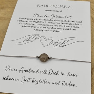 Op de afbeelding: Een grijze draadarmband met een rookkwarts kraal en zilverkleurige accenten. De armband wordt getoond op een kaart met Duitse tekst, waaronder "RAUCHQUARZ" en "Stein der Gelassenheit". De kaart heeft een hart- en handillustratie.