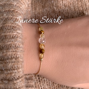 Puede incluir: Un brazalete de cuerda dorada con cuentas marrones, transparentes y amarillas. El brazalete tiene el texto "Innere Stärke".