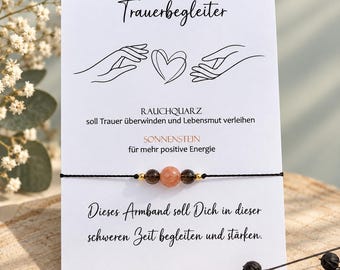 May include: A delicate bracelet featuring a central peach-colored sunstone bead flanked by two dark brown smoky quartz beads and small gold-toned spacers on a thin black cord, presented on a white card with German text and line art illustrations.
